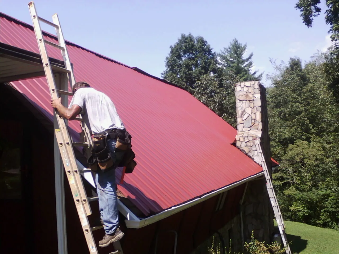 Roofer on a ladder beside a red metal roof and stone chimney — Soffit & Fascia Repair by Roofing Brothers in Forestburgh